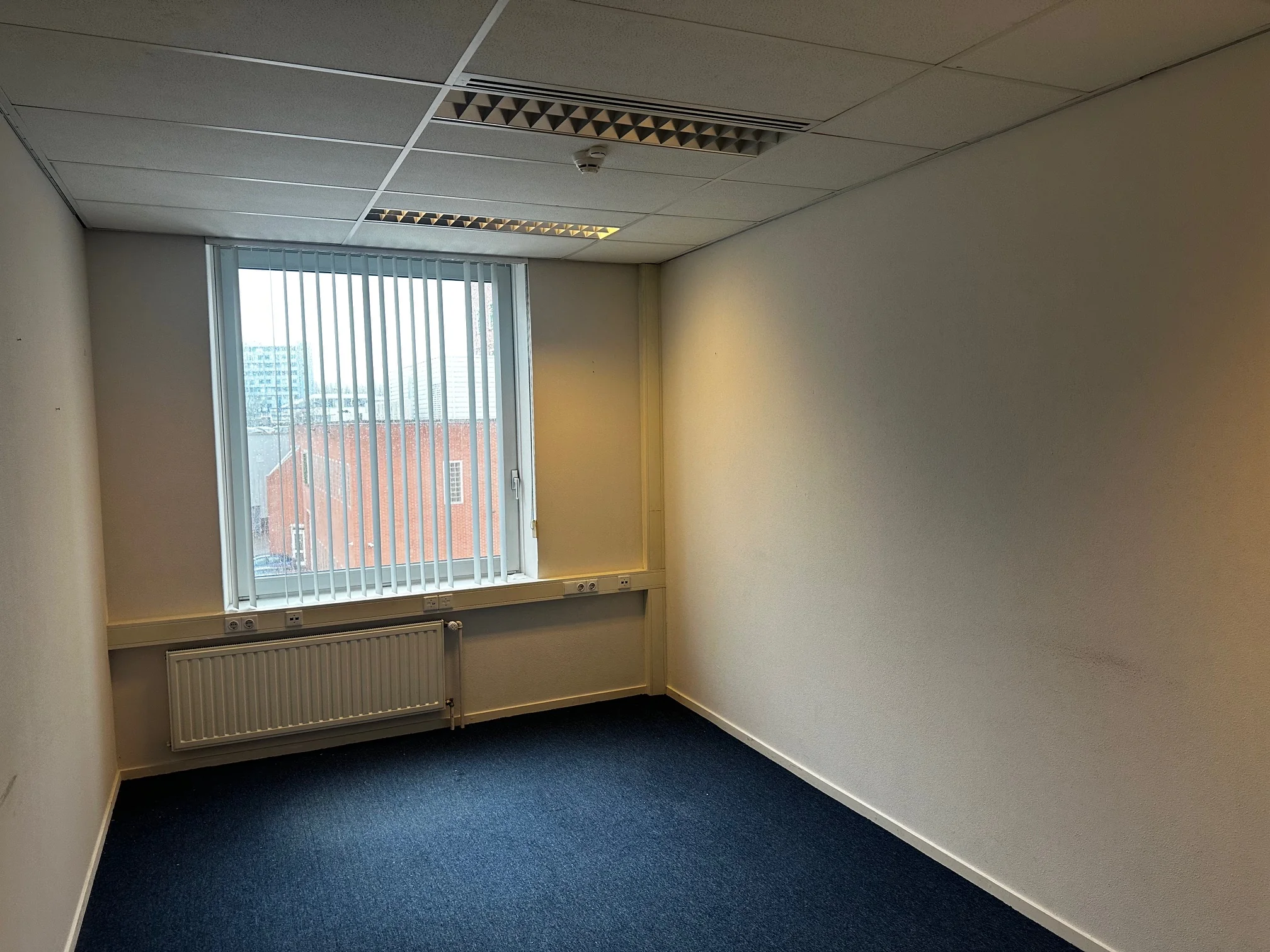 Lege kantoorruimte aan de Van Bleyswijckstraat met blauw tapijt, witte muren en een raam met verticale lamellen.